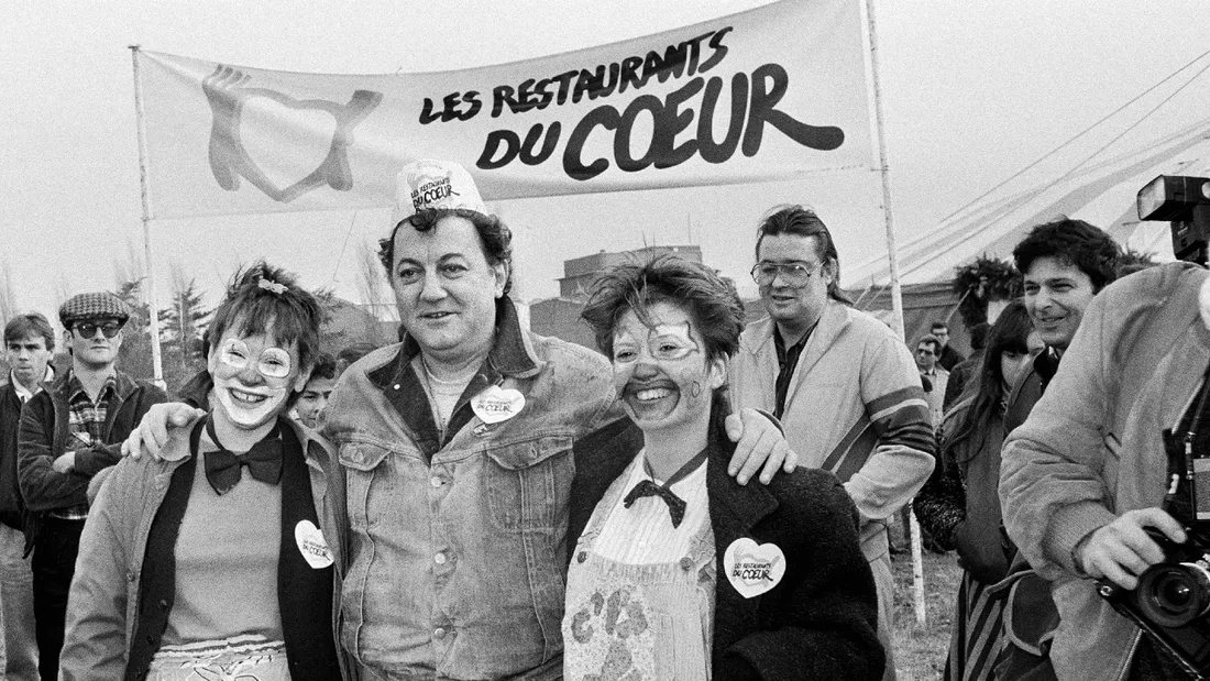 Coluche pose avec deux bénévoles devant l'entrée du Restaurant du Coeur de Gennevilliers en 1985