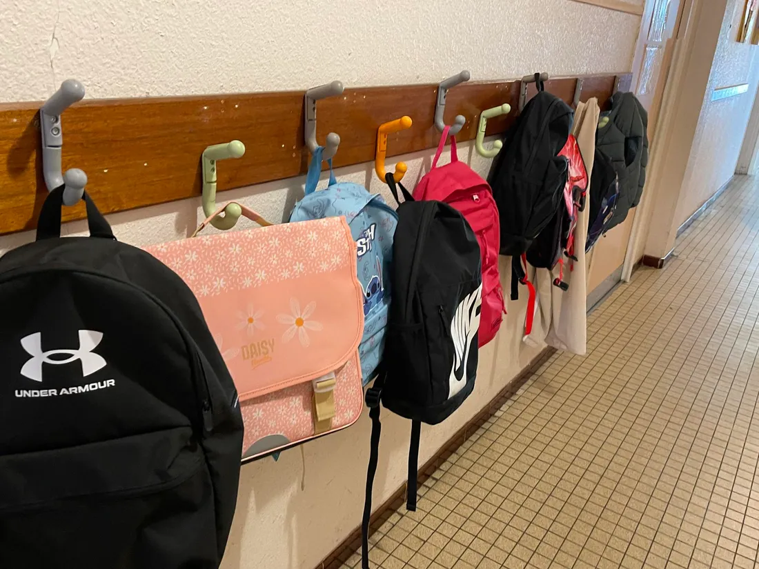 Un couloir dans une école, avec des sacs à dos et des cartables accrochés à des patères.